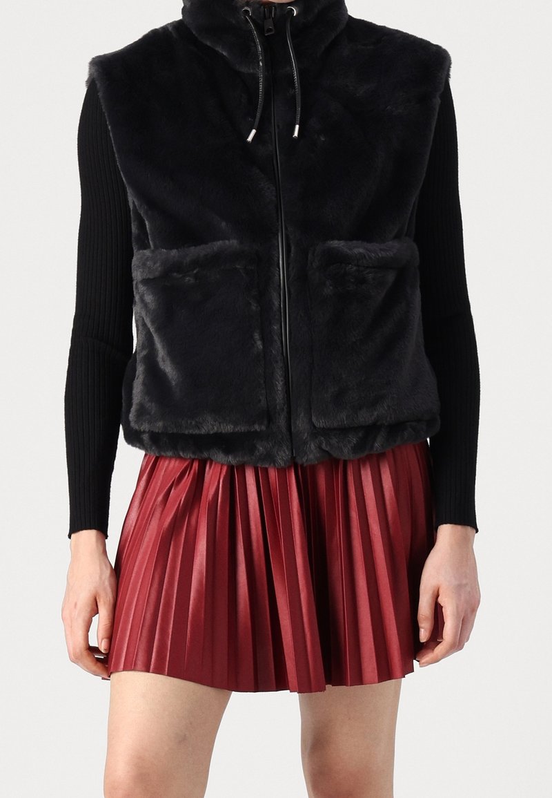 Veste sans manches en fausse fourrure noire zippée avec poches avant, portée sur des manches longues noires et une mini-jupe plissée rouge.