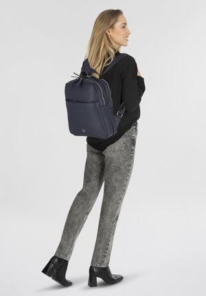 Zaino in pelle blu navy con un design strutturato, dotato di tasche con zip e spallacci regolabili, indossato da un modello su uno sfondo bianco.
