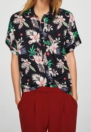 Kvinde iført en sort t-shirt med kort ærme med pink og grøn blomsterprint, stoppet ned i højtaljede røde bukser med hænderne i lommerne.