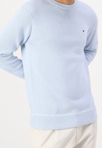 Pull en tricot bleu clair avec un col rond, des poignets et un ourlet côtelés, présentant des motifs texturés subtils et un petit logo accentué.