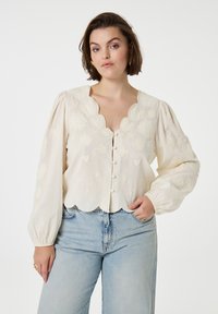 Crème blouse met geborduurde bloemenpatronen, geschulpte zoom en opstaande mouwen. Bevestigd met knopen, gecombineerd met lichtblauwe jeans.
