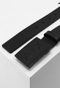 Sort gummibælte med en tekstureret overflade og en rektangulær spænde med indgraverede "AIX" og "ARMANI EXCHANGE" logoer.