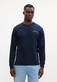 Tumesinine pikakäistega T-särk, mis on valmistatud puuvillast, meeskonnakaelusega, ribakäistega ja rinnal on väike brodiritud logo.