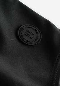 Zwarte stof met een gladde textuur, voorzien van een rond rubberen logo-embleem dat "STAY ACTIVE" en geometrische designelementen bevat.