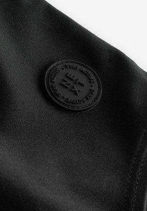 Tissu noir avec une texture lisse présentant un patch en caoutchouc rond embossé avec "RESTEZ ACTIF" et des éléments de design géométriques.