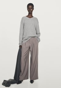 Grauer Strickpullover mit V-Ausschnitt, kombiniert mit hellvioletten plissierten Hosen. Model hält einen dunkelgrauen Schal und trägt schwarze spitze Schuhe.