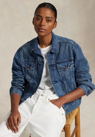 Donna dalla pelle scura e capelli tirati indietro indossa una giacca di jeans blu sopra una camicia bianca e pantaloni bianchi, seduta su una sedia di legno.