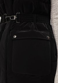 Gilet imbottito nero con texture trapuntata, vita elastica, hardware argentato e una tasca con patta e chiusura a scorrimento. Materiale leggero.
