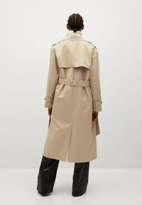 Beżowy płaszcz trenczowy z paskiem w talii, szerokim kołnierzem i detalem na plecach. Posiada długie rękawy i średniej długości krój, noszony na czarne spodnie.