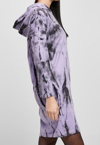 Fioletowa sukienka z kapturem w splot tie-dye z czarnymi wzorami, z długimi rękawami, bocznymi rozcięciami i kapturem. Wykonana z miękkiego materiału, o luźnym kroju.