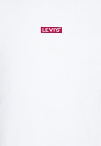 T-shirt blanc à manches courtes en coton, arborant un logo "LEVIs" brodé en rouge sur la poitrine. Texture lisse, coupe classique.