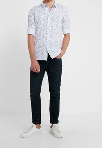 White button-up shirt with a small, colorful pattern, paired with dark jeans and white sneakers. The shirt has rolled sleeves.
