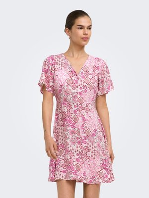 Femme portant une robe fleurie rose à manches courtes et longueur genou avec un col en V, debout devant un fond gris clair uni.