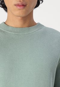 Suéter de malha verde claro com gola redonda canelada, tecido texturizado e design justo, destacando o padrão de malha.