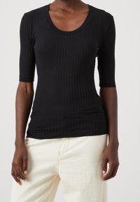 Černá žebrovaná košile s vykrojeným výstřihem a rukávy do lokte, vyrobená z elastického materiálu pro obtáhlý vzhled. Nosit s světle barevnými kalhotami.