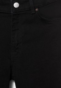 Gros plan sur un jean en denim noir à l’avant avec un bouton argenté et des détails de couture visibles sur la poche et la braguette.