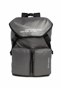 Zaino in nylon grigio con patta pieghevole, spallacci regolabili e due tasche anteriori con zip. Presenta un testo stampato in bianco sulla patta.