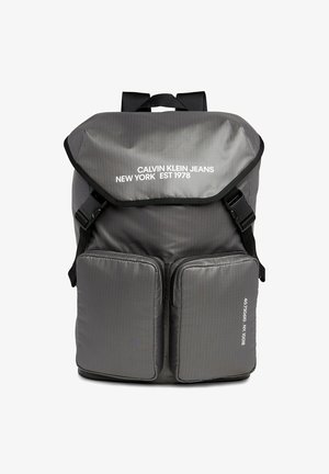 Zaino in nylon grigio con patta pieghevole, spallacci regolabili e due tasche anteriori con zip. Presenta un testo stampato in bianco sulla patta.
