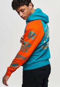 Huvtröja med en ljus turkos kropp och orange ärmar. Har NFL-logotyper och text i kontrasterande färger, medelljus textur.