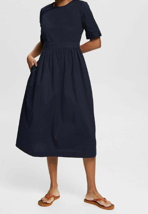 Vrouw draagt een marineblauwe midi-jurk met korte mouwen en zijzakken, gecombineerd met beige slippers, staand tegen een effen achtergrond.