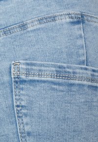 Tecido de denim azul-claro com um bolso traseiro apresentando detalhes de costura visíveis. A textura parece suave com um aspeto clássico de uso.
