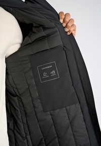 Svart isolerad jacka inuti, med vadderad foder, slät yta och en etikett som indikerar vattenavvisande och vindtåliga egenskaper.