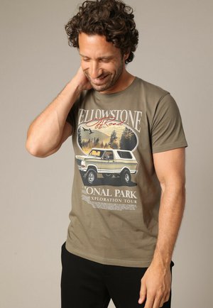 T-shirt en coton vert olive avec un graphisme vintage du parc national de Yellowstone, représentant un camion blanc et une scène naturelle en arrière-plan.