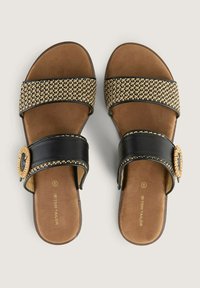 Svarta och bruna slip-on sandaler med breda vävda remmar, texturerad yta och en cirkulär dekorativ detalj på sidan.