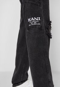 Black denim jeans with a relaxed fit, featuring frayed detailing, white embroidered logo "KANI" on the side pocket, and visible stitching.
