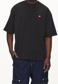 Černé tričko s krátkým rukávem vyrobené z bavlny, s červeným vyšitým logem na levé straně hrudi. Volný střih s jednoduchým kulatým výstřihem.