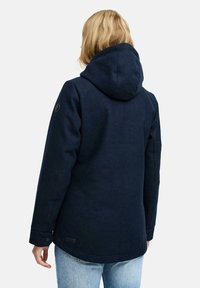 Personne aux cheveux blonds portant une veste à capuche bleu marine et un jean bleu clair, de dos contre un fond clair uni.