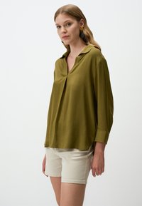 Camicia verde oliva con colletto, taglio morbido, maniche lunghe e spacchi laterali, abbinata a pantaloni corti crema. Texture liscia, design casual.