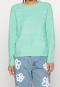 Minzgrüner gestrickter Pullover mit gerippten Bündchen und Saum, ausgestattet mit einem Rundhalsausschnitt und einer leicht lockeren Passform. Kombiniert mit hellblauen Jeanshosen.