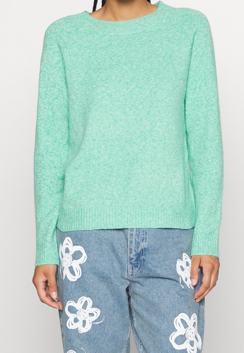 Minzgrüner gestrickter Pullover mit gerippten Bündchen und Saum, ausgestattet mit einem Rundhalsausschnitt und einer leicht lockeren Passform. Kombiniert mit hellblauen Jeanshosen.