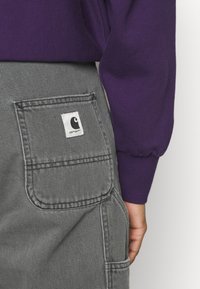 Gray denim overalls with a large utility pocket, featuring a white label with a black logo. Purple long-sleeve shirt partially visible.