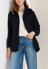 Veste imperméable marine avec une fermeture éclair, une poche avant et une capuche réglable ; associée à un jean large bleu clair.