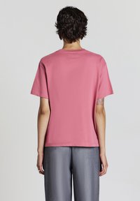 Persona con capelli ricci corti che indossa una maglietta rosa a maniche corte e pantaloni grigi, voltata di spalle rispetto alla fotocamera su uno sfondo bianco.