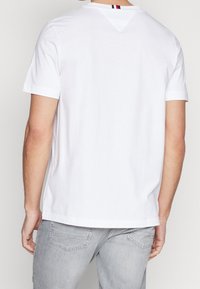 T-shirt bianco in cotone con maniche corte e orlo dritto. La parte posteriore presenta un piccolo accento a righe multicolori vicino al collo.