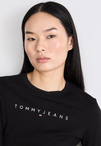 Crna majica dugih rukava s okruglim izrezom. Ima "TOMMYJEANS" logo u bijeloj boji i malu obojenu zastavicu. Glatka tekstura tkanine.