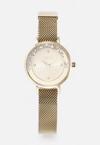 Montre dorée avec un cadran rond ornée d'accentuations scintillantes autour du cadran, bracelet en maille et marqueurs d'heures minimalistes.