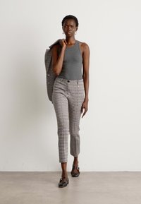 Graues Tanktop kombiniert mit gemusterten Hosen, die eine subtile Textur aufweisen. Das Model hält ein Blazer und trägt schwarze Ballerinas. Einfaches, maßgeschneidertes Design.