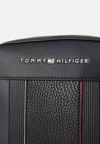 Černá texturovaná kožená peněženka s vertikálními drážkami, se stříbrným logem "TOMMY HILFIGER" a červeným akcentním pruhem.