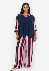 Suéter de punto azul marino de gran tamaño con escote en V, superpuesto a una blusa de rayas multicolores y pantalones de rayas holgados en rosa, azul marino y burdeos.