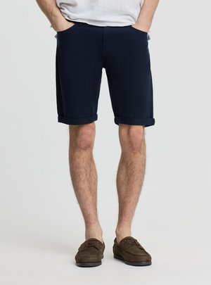 Uomo che indossa pantaloncini blu navy risvoltati, una camicia bianca e mocassini marroni, in piedi con le mani in tasca su uno sfondo semplice.