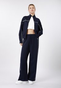 Ensemble de survêtement bleu marine comprenant une veste zippée et un pantalon à jambes larges, agrémenté de passepoils blancs. Associé à des baskets blanches sur un fond neutre.