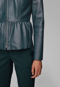 Teal leather jacket with a fitted design, pleated waist, and zip closure. Long sleeves with a smooth texture. Paired with dark pants.