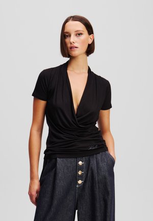Black short-sleeve top with a deep V-neck and draped design. Features a gathered texture at the front and a logo accent on the hem.