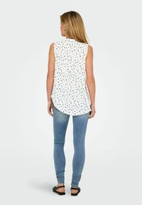 Ærmeløs hvid top med blåt blomsterprint, buet kant, parret med tætsiddende lyseblå jeans og sorte sandaler.