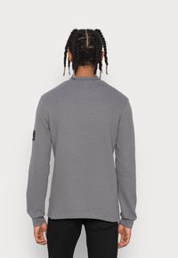 Grå långärmad skjorta i textil med struktur, med ribbade ärmslut och en etikett på vänster ärm. Bakåtsidan visar en enkel design.