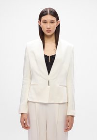 Blazer branco ajustado com lapela xale, fecho frontal com botões e dois bolsos laterais. Textura suave com um corte estruturado.
